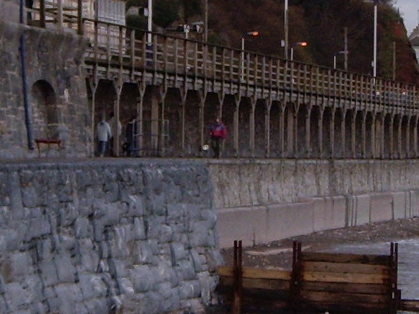 The image shows a stone sea wall and a railing extending along a coastal walkway. There are a few people walking and standing near the railing. The photograph appears during dusk with lighting along the walkway illuminating the scene. Due to the nature of the image, it seems to demonstrate the camera's low-light photography capabilities, possibly taken with a Pentax Optio T20 camera. The image has a moderate level of details with noticeable softness, suggesting either motion blur or a limited performance in low-light conditions.