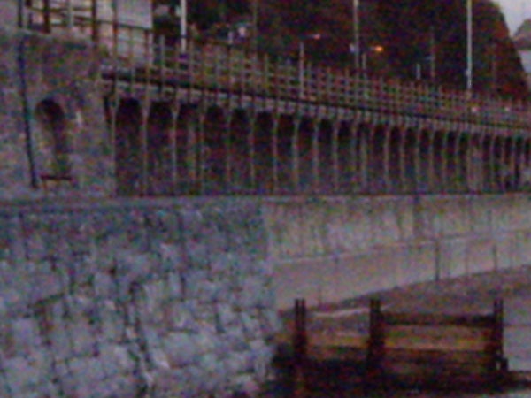 Low-light photograph showing a fence alongside a seawall and a pier taken with Pentax Optio T20, demonstrating noise and detail at high ISO settings.