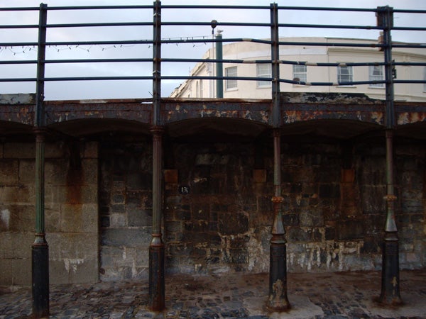 Photograph taken with a Pentax Optio T20 showing an old stone structure with arched openings and metal poles, with a building visible in the background and birds perched on electrical wires above.