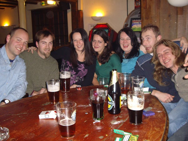 A group of seven people smiling and enjoying drinks together around a wooden table at a pub.