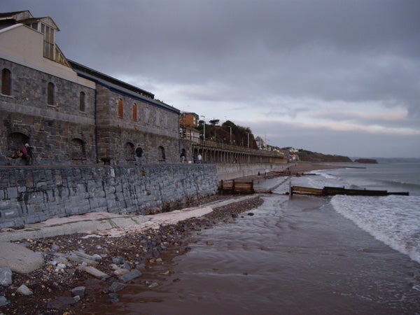 Photograph taken with Pentax Optio T20 camera showing a cloudy day at the beach with foamy waves reaching the shore, a stone sea wall on the left, and a line of buildings and a promenade extending into the distance.