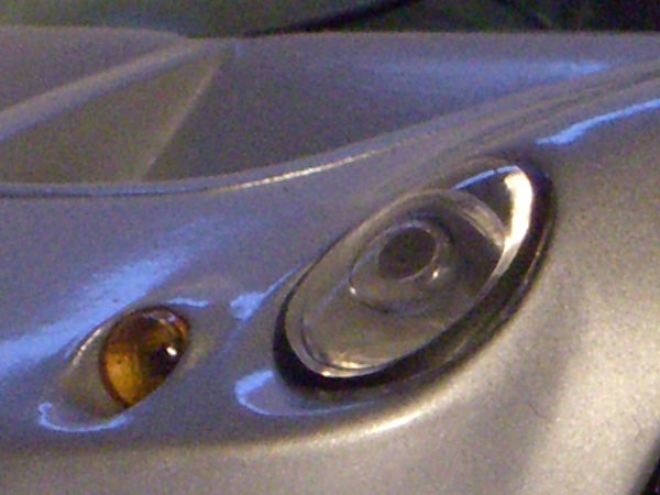 Close-up photo of a grey surface with a clear oval plastic cover, potentially a part of an electronic device, with a smaller amber-colored detail beside it.