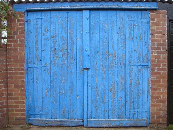 A weathered blue wooden double door of a garage with peeling paint and visible brickwork on the side.