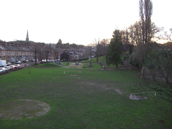 A photograph taken with a Casio Exilim EX-S770 showcasing a park at dusk, with visible green grass, bare trees, a playground, and a row of parked cars along a street, with houses and a church spire in the background.