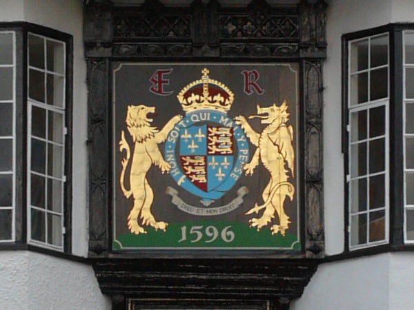Photo of a historical crest with a crown at the top, flanked by two golden lions, against a black timbered building facade. The crest features a shield with a blue and red design, Latin text, and the date 1596 displayed prominently at the bottom.