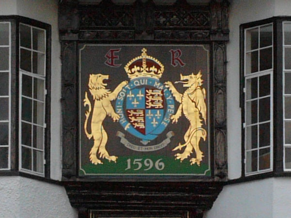 The image is a detailed photograph of an elaborate coat of arms featuring two lions and a shield with various heraldic symbols, set against a building with decorative timber framing. The coat of arms includes a crown at the top and the date 1596 prominently displayed at the bottom.