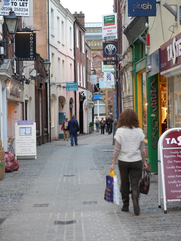 Photograph taken with a Panasonic DMC-L1 Digital SLR depicting a cobblestone street scene with pedestrians and shops, demonstrating the camera's depth of field and color rendition in a real-world setting.