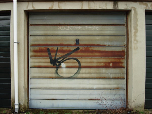 The image displays a rusty metal garage door with graffiti scrawled across it.