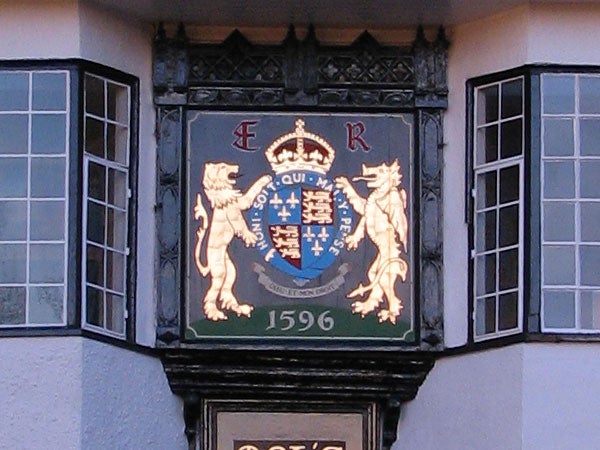 The image displays an ornate, heraldic coat of arms sign with the date 1596, situated between two windows. The coat of arms is adorned with a crown at the top and flanked by two golden standing lion figures.