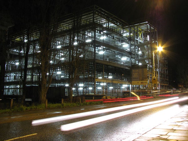 A nighttime long-exposure photograph taken with the Canon PowerShot S3 IS, capturing the light trails of a passing car in front of a multi-story construction site illuminated by artificial lights.