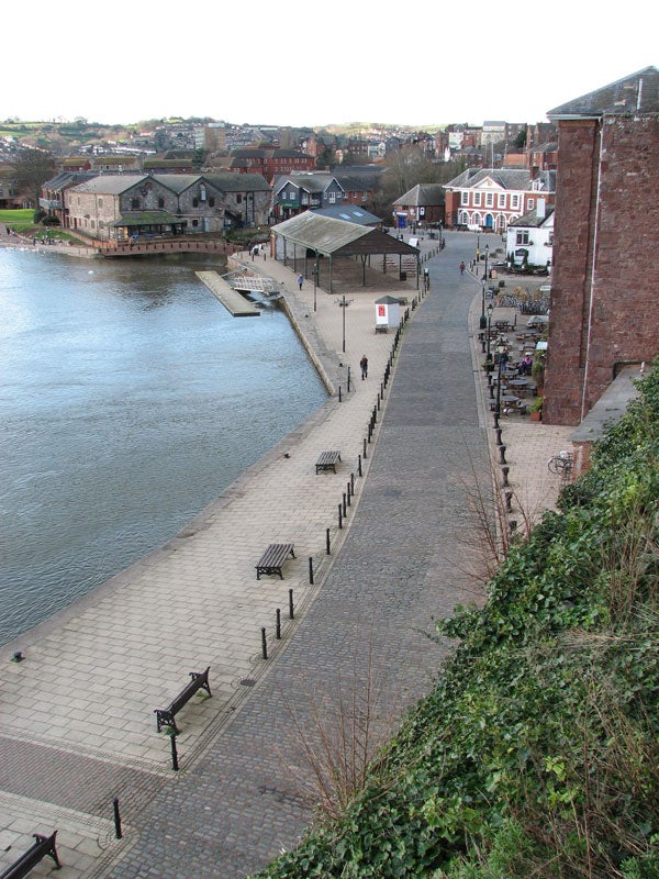 High-angle view over a riverside walkway with benches, leading to a small pavilion by the water's edge, with buildings and a red brick wall on the right, and vegetation on the left.