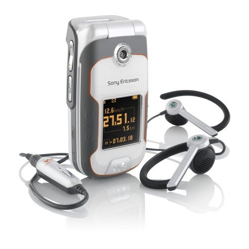Sony Ericsson W710i mobile phone displayed with its flip open, showcasing screen and keypad, alongside a set of sports earphones and a car charger, all arranged on a reflective white surface.