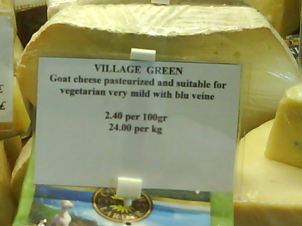 The image shows a label for Village Green goat cheese, with details about it being pasteurized and suitable for vegetarians, with a mild taste and blue vein. The pricing is listed as 2.40 per 100 grams and 24.00 per kilogram. There is a blurry background that seems to be a cheese display. The image has no direct relation to the Sony Ericsson W710i.