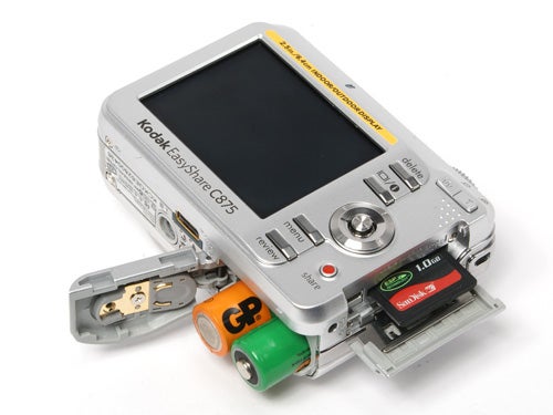 Kodak EasyShare C875 digital camera with the battery compartment open showing two AA batteries and an SD memory card inserted.