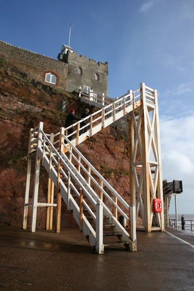 A clear, well-lit photo depicting a set of white wooden stairs leading to a historic building perched atop a cliff with a backdrop of a cloudy blue sky. The image also shows a part of the beach and a figure in the distance, illustrating the camera's capability to capture detail and color contrast in natural lighting.