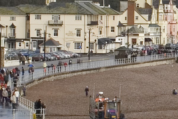 Photograph taken with a Canon EOS 400D showcasing its zoom capability, featuring a seaside promenade scene with pedestrians, a pebble beach, buildings in the background, and a tractor in the foreground.