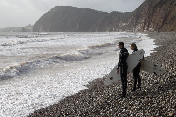 Two surfers standing on a pebble beach with surfboards, looking out at the rough sea with a cliff in the background, likely taken with a Canon EOS 400D camera.