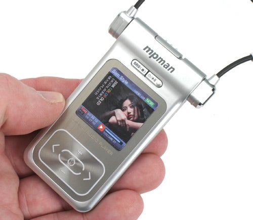 A hand holding an MPMan MP3 player with a color screen displaying a music track and playback controls.