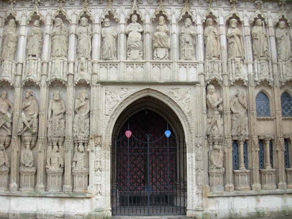 The image displays an intricate cathedral entrance with detailed stone carvings of figures and ornate gothic architecture. A large wooden door is at the center, flanked by stone statues within niches.