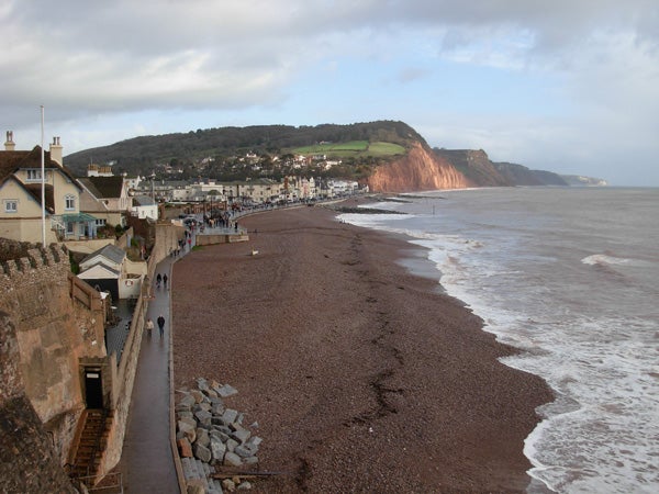 A Nikon CoolPix L5 photo showcasing a pebble beach with a promenade to the left, houses lining the street, cliffs in the distance, and a cloudy sky.
