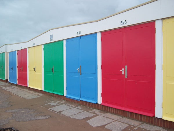 A row of colorful beach huts with closed doors in red, blue, green, yellow, and white, captured under an overcast sky.