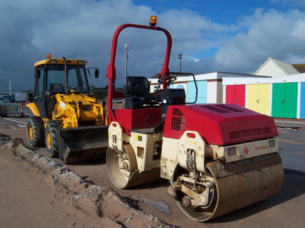 Construction vehicles on road work site with colorful beach huts in the background.