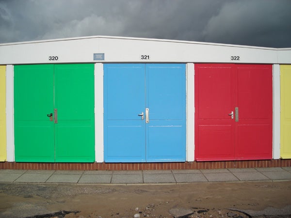 Three colorful beach huts with closed doors, numbered 320 to 322, in green, blue, and red respectively, under a cloudy sky.