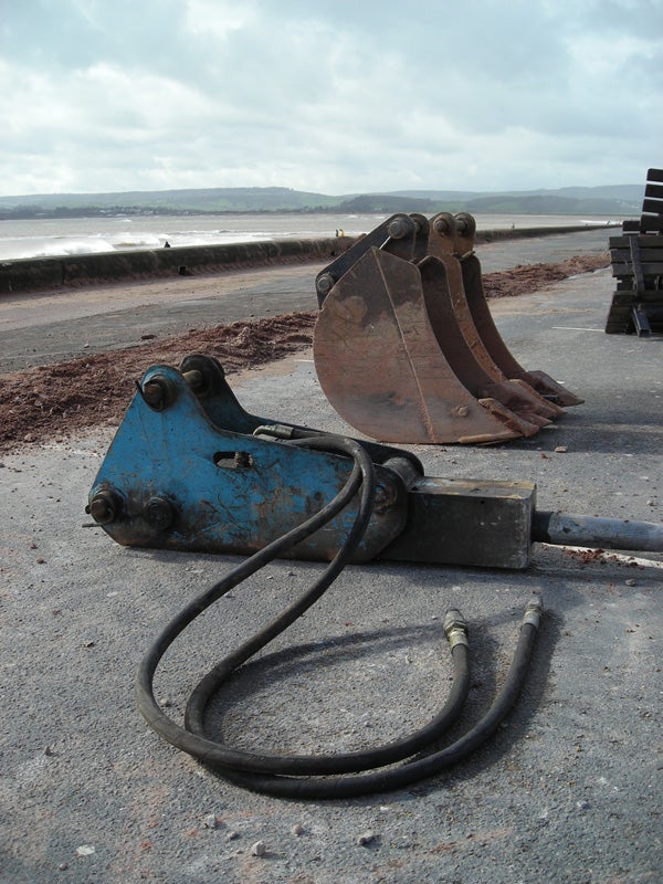 A Nikon CoolPix L6 photo of a blue excavator bucket attachment resting on a paved surface with a beach and the sea in the background under an overcast sky.