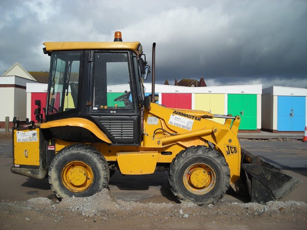 A yellow JCB backhoe loader parked on a construction site with colorful beach huts in the background and a cloudy sky above.