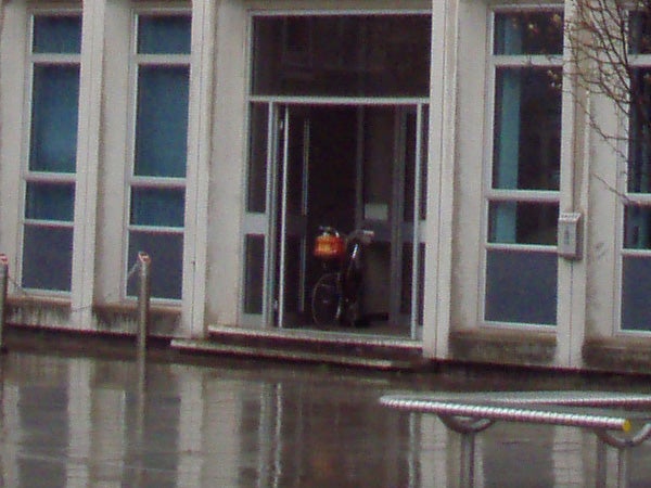 Orange bike parked under a covered entrance of a building with blue windows on a rainy day.