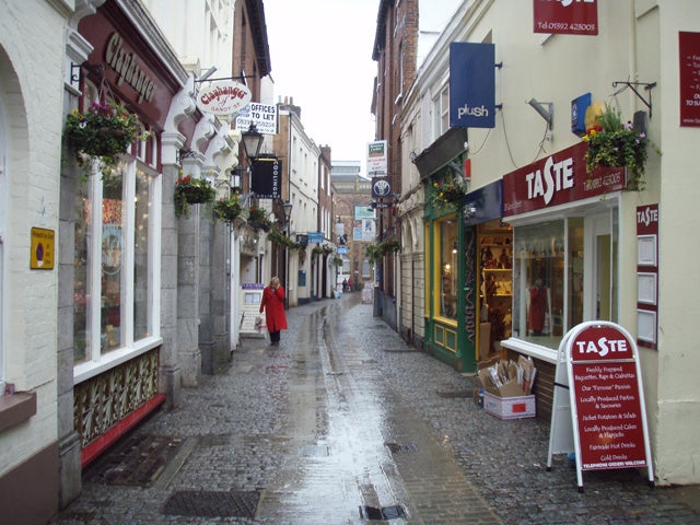 A cobblestone street with wet ground after rain, featuring old European architecture, various shop signs, and a person in a red coat walking in the distance.