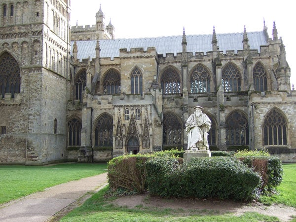 The image shows a statue located in front of an old, gothic-style church with intricate architectural details. The church has numerous arched windows and is adorned with Gothic decorations. A well-maintained lawn surrounds the statue, and a neatly trimmed hedge encircles its base. The weather appears to be clear and sunny, which highlights the sharpness and color fidelity of the photo, likely demonstrating the image quality achievable with the Fujifilm FinePix F20 camera.