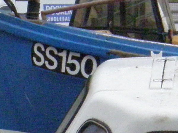 Close-up photo demonstrating the zoom capabilities of the Olympus SP-550UZ camera, showing a blue boat with the registration 'SS150' on the side, partially obscured by the white roof of another boat.