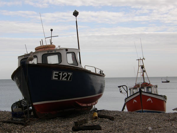 A photograph taken with an Olympus SP-550UZ showing two boats on a shingle beach with the ocean in the background and cloudy skies above.