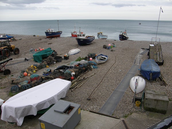 The image shows a pebbled beach with fishing boats, assorted boating equipment, and fishing nets scattered throughout. The sea is visible in the background, under a cloudy sky. The photo is likely taken from an elevated vantage point capturing a broad view of the scene.