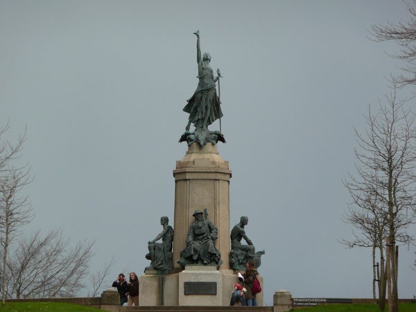 A bronze statue monument featuring multiple figures on a stone pedestal with a clear sky in the background, with people sitting and standing around its base.