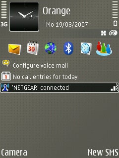 Nokia E65 smartphone home screen showing date and time, signal strength, carrier label 'Orange', and various application icons including email, web browser, and connectivity status indicating 'NETGEAR' connected. Options for 'Camera' and 'New SMS' are displayed at the bottom.
