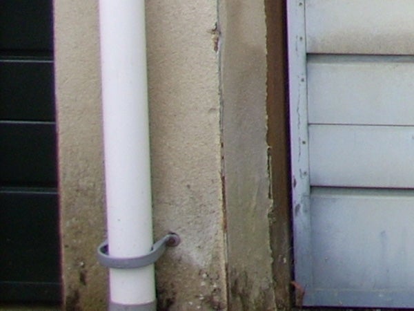 A close-up photo showing a white drainpipe attached to a beige wall next to a window with closed shutters, potentially demonstrating the detail and color capture quality of the Nikon CoolPix L10 camera.