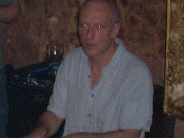 Man sitting indoors with a glass on the table in front of him, showing red-eye effect typically associated with flash photography. The lighting is dim and the image quality suggests a low-light capture scenario.