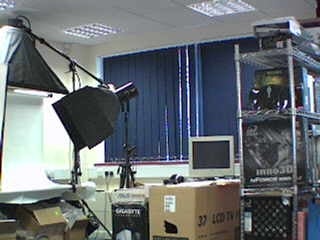 A photo taken from the TRENDnet TV-IP201W IP Camera showing an office environment with various electronic equipment, lighting setups, and boxed items.