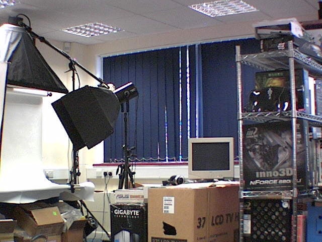 Image showing an office with technological equipment, including computer components and boxes for Gigabyte and Asus products, a CRT monitor, and photographic lighting equipment. The image appears to be captured through a TRENDnet TV-IP201W IP Camera, illustrating the camera's visual field and image quality within an indoor setting.