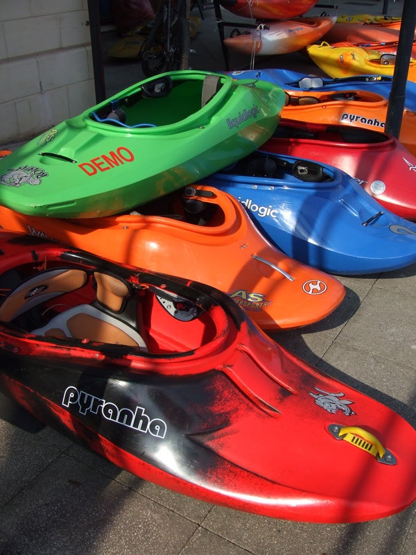 A vividly colorful array of kayaks neatly arranged, showcasing different models and sizes with clearly visible brand names and designs, photographed under bright natural lighting.