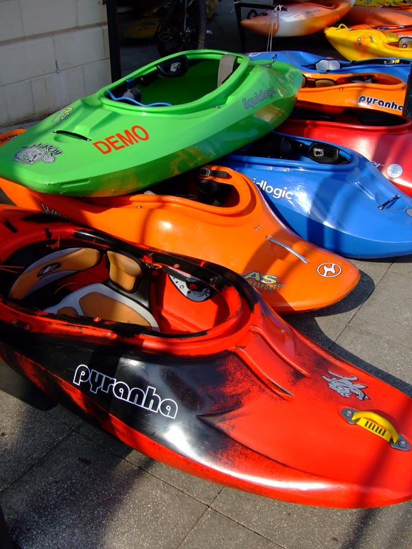 The image shows a stack of colorful kayaks in red, blue, green, and yellow, with brand names such as Pyranha and Logic visible on their surfaces, arranged outdoors for display or sale.