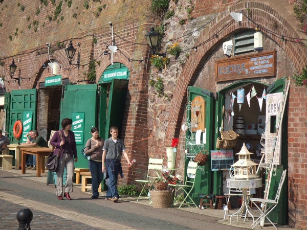 Photograph showing a quaint street scene with people walking past shops built into archways under a brick bridge, with various items like baskets and a white birdcage on display outside the shops.