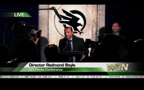 Screenshot from the video game Command and Conquer 3: Tiberium Wars showing a character known as Director Redmond Boyle at a GDI press conference with journalists and cameras in the foreground, and a GDI logo in the background.