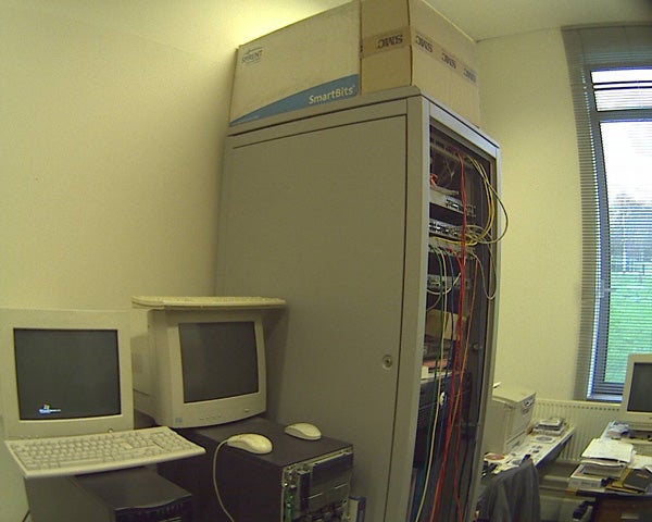 View of a server room showing a large network equipment cabinet labeled 'SmartBits', with colorful wires and cables, alongside an old-style CRT monitor on a desk, captured by the Axis 207MW Wireless Network Camera.