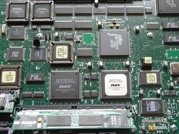 Close-up view of a green circuit board with various integrated circuits, chips, and electronic components, prominently featuring several ALTERA FLEX chips.