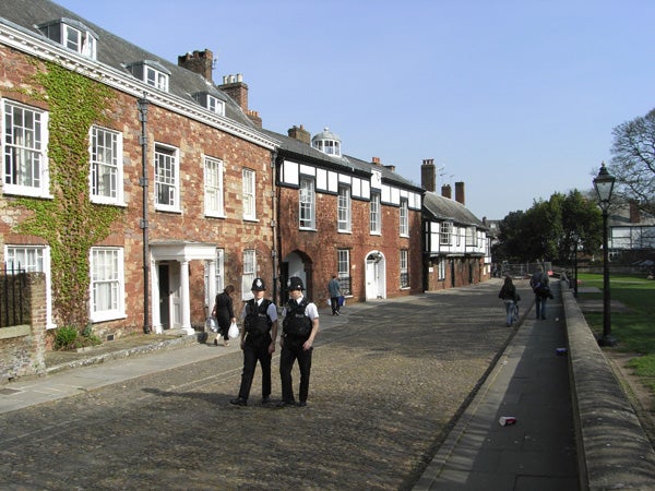 Image of a sunny street scene captured with a Ricoh Caplio R6 camera, featuring uniformed police officers in the foreground conversing, historic brick buildings with white windows on the side, and a few pedestrians in the distance walking on the pavement.