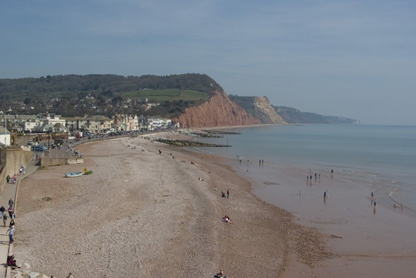 A beachscape shot with Samsung GX-10 showing a clear view of a pebble beach with people, seafront buildings, and tall cliffs on a sunny day.