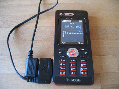 Sony Ericsson W880i mobile phone in black and orange, connected to a charger, displaying the T-Mobile ringtone screen on a wooden surface.